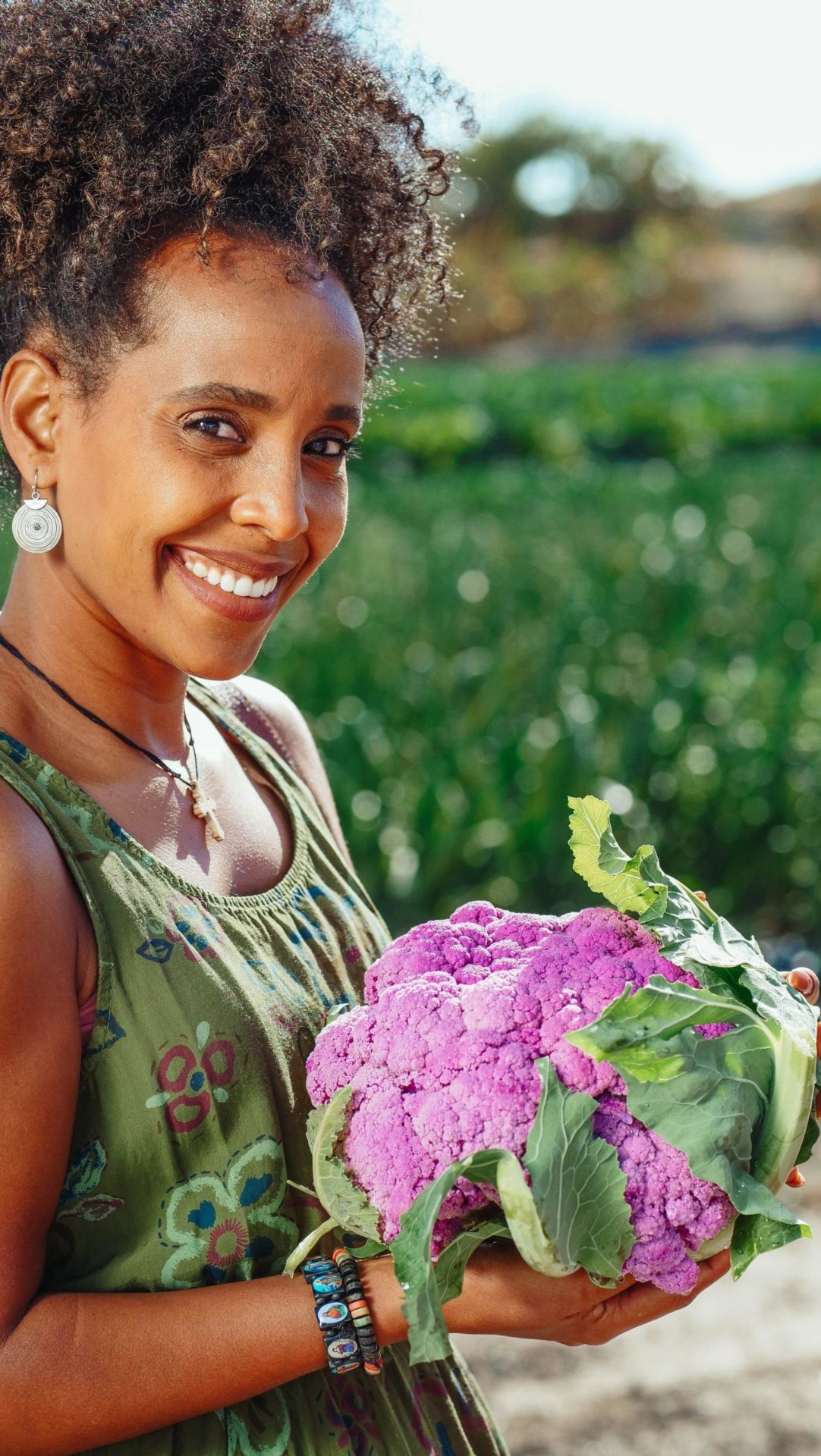 Smiling Woman in Green Top Holding a purple Cauliflower in the fresh air overlooking her garden