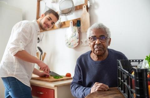 An Elderly Man Sitting while Wearing Eyeglasses while a Volunteer prepares him a meal