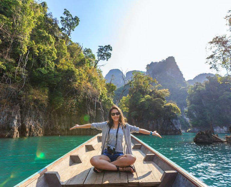 Smiling woman on a boat on holiday with her arms out-stretched in front of some cliffs