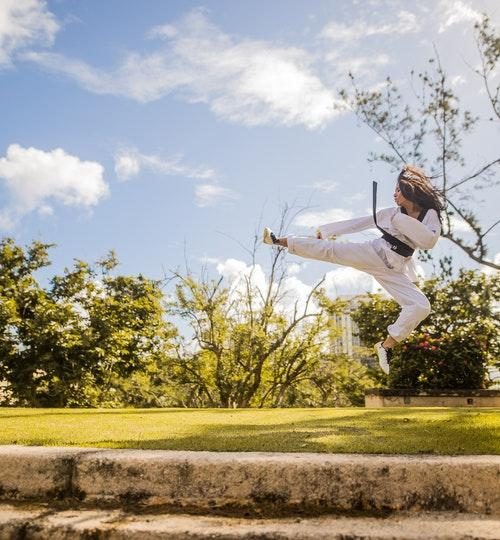 Woman Wearing White Karati G Under Blue Sky doing a karate kick in the air over grass at the park