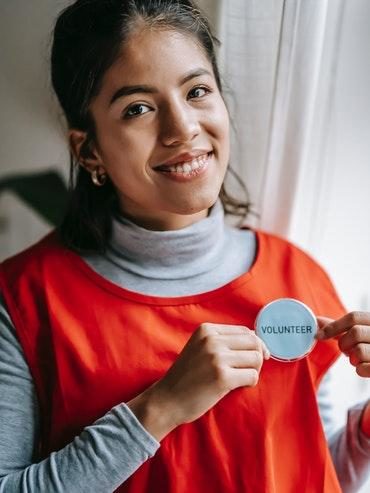 A smiling lady wearing an orange overall and proudly showing off her Volunteer badge boosting her own well-being by helping others selflessly